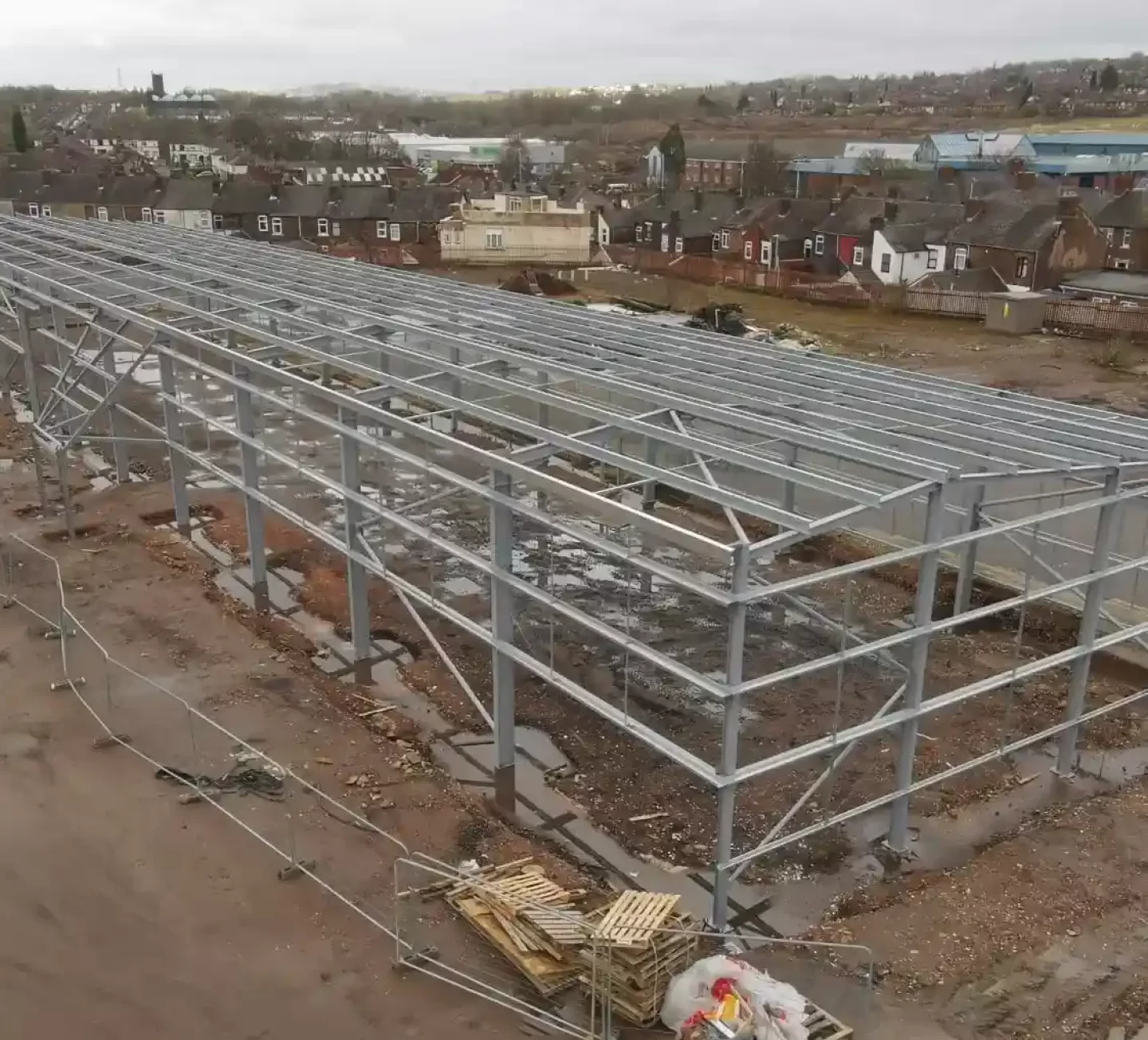 Large-scale steel frame structure for a commercial or industrial warehouse under construction, with residential buildings visible in the background.